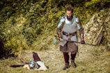 A bearded man in medieval armor stands in a forest clearing, holding a large axe. He wears chainmail and metal shoulder guards, along with a leather belt and gloves. On the ground lies another armored figure, seemingly lifeless, surrounded by grass and foliage.