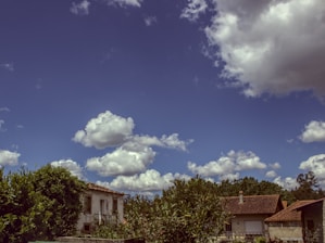 A rural scene featuring two houses with tiled roofs surrounded by lush greenery, under a blue sky dotted with large, fluffy white clouds.