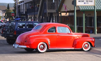 A bright red vintage car is parked in a parking lot beside a navy blue SUV with a bicycle mounted on its back. The setting includes a motel or roadside building, evident from the architecture and a sign offering amenities like 'Micro Fridge' and 'WiFi'. An American flag is visible in the background.