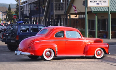 A bright red vintage car is parked in a parking lot beside a navy blue SUV with a bicycle mounted on its back. The setting includes a motel or roadside building, evident from the architecture and a sign offering amenities like 'Micro Fridge' and 'WiFi'. An American flag is visible in the background.
