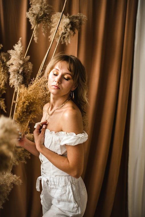 A serene, stylish woman confidently posing in a softly lit room filled with plants and elegant decor.