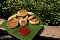 A serving of golden-brown fried patties is neatly arranged on a bright green banana leaf. A dollop of red sauce is placed beside the patties. The setup is outdoors, with abundant green foliage in the background and laid upon a wooden surface.