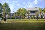 A group of people are playing cricket on a well-maintained lawn in front of a modern house. The setting is surrounded by lush greenery, including palm trees and bushes. There are a few scattered clouds in a blue sky, contributing to a sunny atmosphere. One person is preparing to bowl, and others are positioned around the field, with a white chair and a stool seen on the grass.