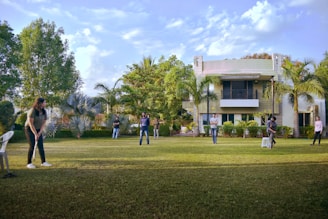 A group of people are playing cricket on a well-maintained lawn in front of a modern house. The setting is surrounded by lush greenery, including palm trees and bushes. There are a few scattered clouds in a blue sky, contributing to a sunny atmosphere. One person is preparing to bowl, and others are positioned around the field, with a white chair and a stool seen on the grass.