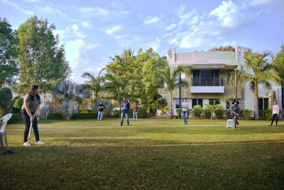 A group of people are playing cricket on a well-maintained lawn in front of a modern house. The setting is surrounded by lush greenery, including palm trees and bushes. There are a few scattered clouds in a blue sky, contributing to a sunny atmosphere. One person is preparing to bowl, and others are positioned around the field, with a white chair and a stool seen on the grass.