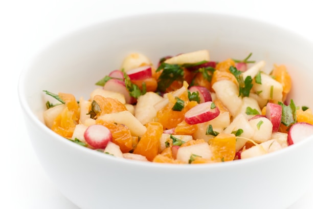 A white bowl filled with a colorful salad consisting of diced fruits and vegetables such as oranges, radishes, apples, and fresh herbs.