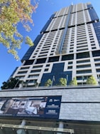 A tall, modern skyscraper with a sleek, geometric design features numerous windows and balconies. The foreground displays a sign advertising luxury penthouse suites with information on sales and rentals. Several small trees and flagpoles are lined up on the terrace, adding a touch of greenery to the urban setting.