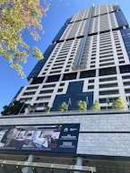 A tall, modern skyscraper with a sleek, geometric design features numerous windows and balconies. The foreground displays a sign advertising luxury penthouse suites with information on sales and rentals. Several small trees and flagpoles are lined up on the terrace, adding a touch of greenery to the urban setting.