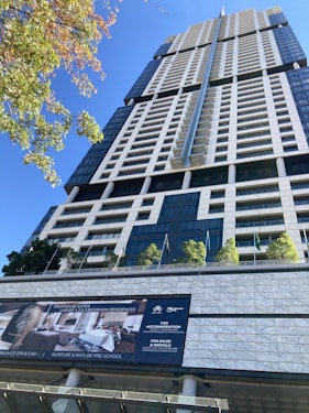 A tall, modern skyscraper with a sleek, geometric design features numerous windows and balconies. The foreground displays a sign advertising luxury penthouse suites with information on sales and rentals. Several small trees and flagpoles are lined up on the terrace, adding a touch of greenery to the urban setting.