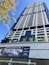 A tall, modern skyscraper with a sleek, geometric design features numerous windows and balconies. The foreground displays a sign advertising luxury penthouse suites with information on sales and rentals. Several small trees and flagpoles are lined up on the terrace, adding a touch of greenery to the urban setting.