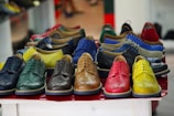 A collection of colorful leather dress shoes is arranged on a tabletop. The shoes come in various vibrant colors, including green, yellow, brown, red, and blue. They feature classic brogue detailing and are neatly aligned in rows, creating an eye-catching display.