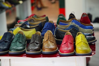 A collection of colorful leather dress shoes is arranged on a tabletop. The shoes come in various vibrant colors, including green, yellow, brown, red, and blue. They feature classic brogue detailing and are neatly aligned in rows, creating an eye-catching display.