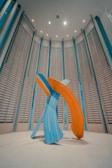 A modern art sculpture consisting of large, abstract brush strokes in vibrant blue and orange hues. The sculpture is set in a spacious room with high ceilings and vertically aligned, louvered wall panels. The lighting is soft, casting gentle shadows on the floor.