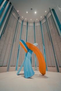 A modern art sculpture consisting of large, abstract brush strokes in vibrant blue and orange hues. The sculpture is set in a spacious room with high ceilings and vertically aligned, louvered wall panels. The lighting is soft, casting gentle shadows on the floor.