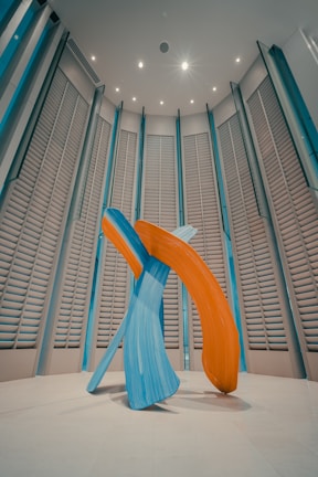 A modern art sculpture consisting of large, abstract brush strokes in vibrant blue and orange hues. The sculpture is set in a spacious room with high ceilings and vertically aligned, louvered wall panels. The lighting is soft, casting gentle shadows on the floor.