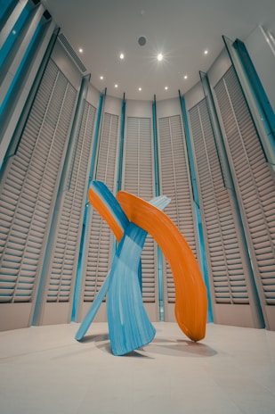 A modern art sculpture consisting of large, abstract brush strokes in vibrant blue and orange hues. The sculpture is set in a spacious room with high ceilings and vertically aligned, louvered wall panels. The lighting is soft, casting gentle shadows on the floor.