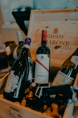 A wooden box filled with several bottles of wine and scattered corks. The focus is on a variety of wine bottles, each with distinct labels and corks around them. The box has printed text and an elegant font indicating a brand or type of wine.