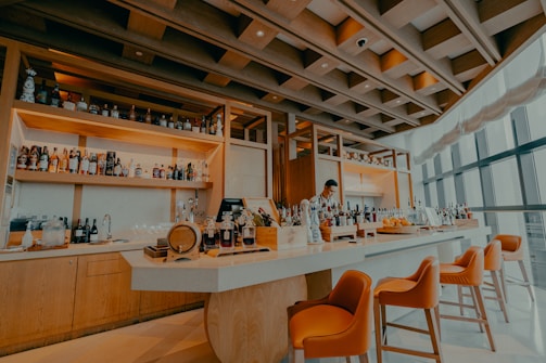 A modern bar with a wooden ceiling and shelves displaying a variety of liquor bottles. A bartender stands behind the white bar counter which is adorned with decorative bottles and a small wooden barrel. Orange bar stools are neatly positioned along the counter, and large windows allow natural light to fill the space.