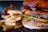 A close-up of a juicy cheeseburger with layers of bacon, cheddar cheese, pickles, lettuce, and tomato. The burger is set in a toasted bun. Next to the burger is a generous portion of thick-cut French fries with a slightly golden crispy texture.