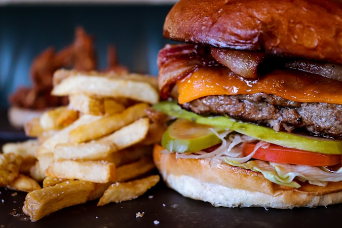 A close-up of a juicy cheeseburger with layers of bacon, cheddar cheese, pickles, lettuce, and tomato. The burger is set in a toasted bun. Next to the burger is a generous portion of thick-cut French fries with a slightly golden crispy texture.