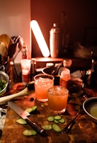 A dimly lit kitchen counter with two cocktails garnished with cucumber slices. The cocktails are surrounded by a variety of bar tools and ingredients, including a wooden cutting board, a knife, and a shaker. There is an orange glow from a light, creating a warm ambiance.