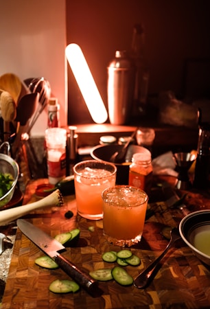 A dimly lit kitchen counter with two cocktails garnished with cucumber slices. The cocktails are surrounded by a variety of bar tools and ingredients, including a wooden cutting board, a knife, and a shaker. There is an orange glow from a light, creating a warm ambiance.