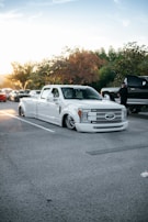 A customized white truck with a low suspension is parked in a parking lot. The surroundings include a few other vehicles and some trees with autumn-colored leaves. A person is standing near a black vehicle, and the sun is setting in the background, casting a warm glow over the scene.