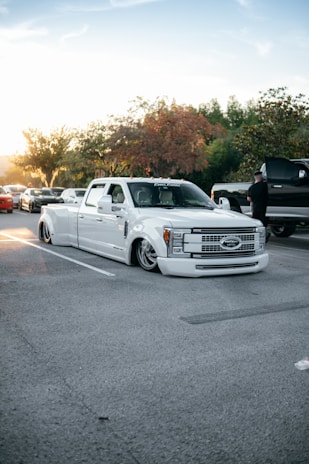 A customized white truck with a low suspension is parked in a parking lot. The surroundings include a few other vehicles and some trees with autumn-colored leaves. A person is standing near a black vehicle, and the sun is setting in the background, casting a warm glow over the scene.