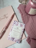 A flatlay image featuring a pink-themed scene with a notepad inscribed with the word 'breathe', a glittery pink pen, a packet of Greenleaf Prosecco Plum Scented Envelope Sachet, a pink candle, and a pink cloth. The items are arranged on a marble surface, and the elements convey a soothing and relaxing atmosphere.