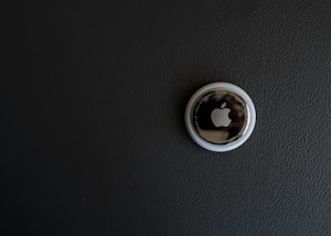 A single Apple AirTag is placed on a dark textured surface. The AirTag, a small circular device, has a glossy white outer ring and a metallic center with the Apple logo prominently displayed.