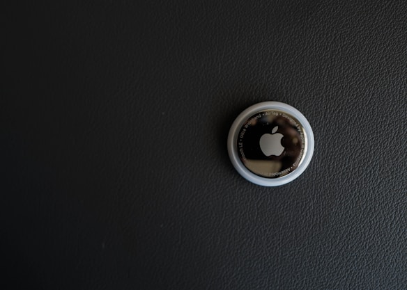 A single Apple AirTag is placed on a dark textured surface. The AirTag, a small circular device, has a glossy white outer ring and a metallic center with the Apple logo prominently displayed.