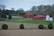 A rural landscape featuring a bright red barn and outbuildings located on a gently sloping hill. In the foreground, there is a field with neatly spaced trees, and in the background, a line of trees with early spring foliage. Silver silos are positioned to the right of the barn, adding a reflective contrast to the scene.