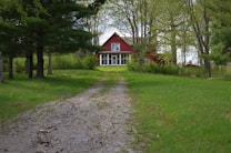 A rustic red house with a white-trimmed porch is nestled among tall green trees. A gravel pathway leads up to the house, surrounded by a vibrant green lawn. The setting is serene and somewhat secluded, evoking a sense of calm and nature.