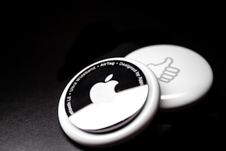 Two white circular devices are placed against a dark background. The one in the foreground features an Apple logo and text around its edge, including phrases like 'Bluetooth LE' and 'Ultra Wideband.' The one in the background shows a thumbs-up symbol.