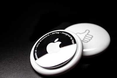 Two white circular devices are placed against a dark background. The one in the foreground features an Apple logo and text around its edge, including phrases like 'Bluetooth LE' and 'Ultra Wideband.' The one in the background shows a thumbs-up symbol.
