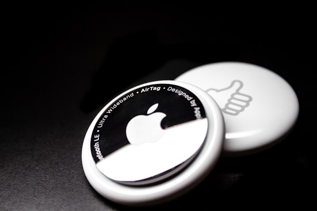Two white circular devices are placed against a dark background. The one in the foreground features an Apple logo and text around its edge, including phrases like 'Bluetooth LE' and 'Ultra Wideband.' The one in the background shows a thumbs-up symbol.