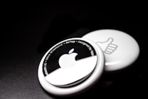 Two white circular devices are placed against a dark background. The one in the foreground features an Apple logo and text around its edge, including phrases like 'Bluetooth LE' and 'Ultra Wideband.' The one in the background shows a thumbs-up symbol.