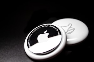 Two white circular devices are placed against a dark background. The one in the foreground features an Apple logo and text around its edge, including phrases like 'Bluetooth LE' and 'Ultra Wideband.' The one in the background shows a thumbs-up symbol.