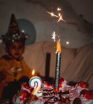 A brightly decorated cake with a large number two candle and a lit sparkler on top. The cake is adorned with strawberries and chocolate shavings. In the background, slightly out of focus, is a child wearing a party hat.