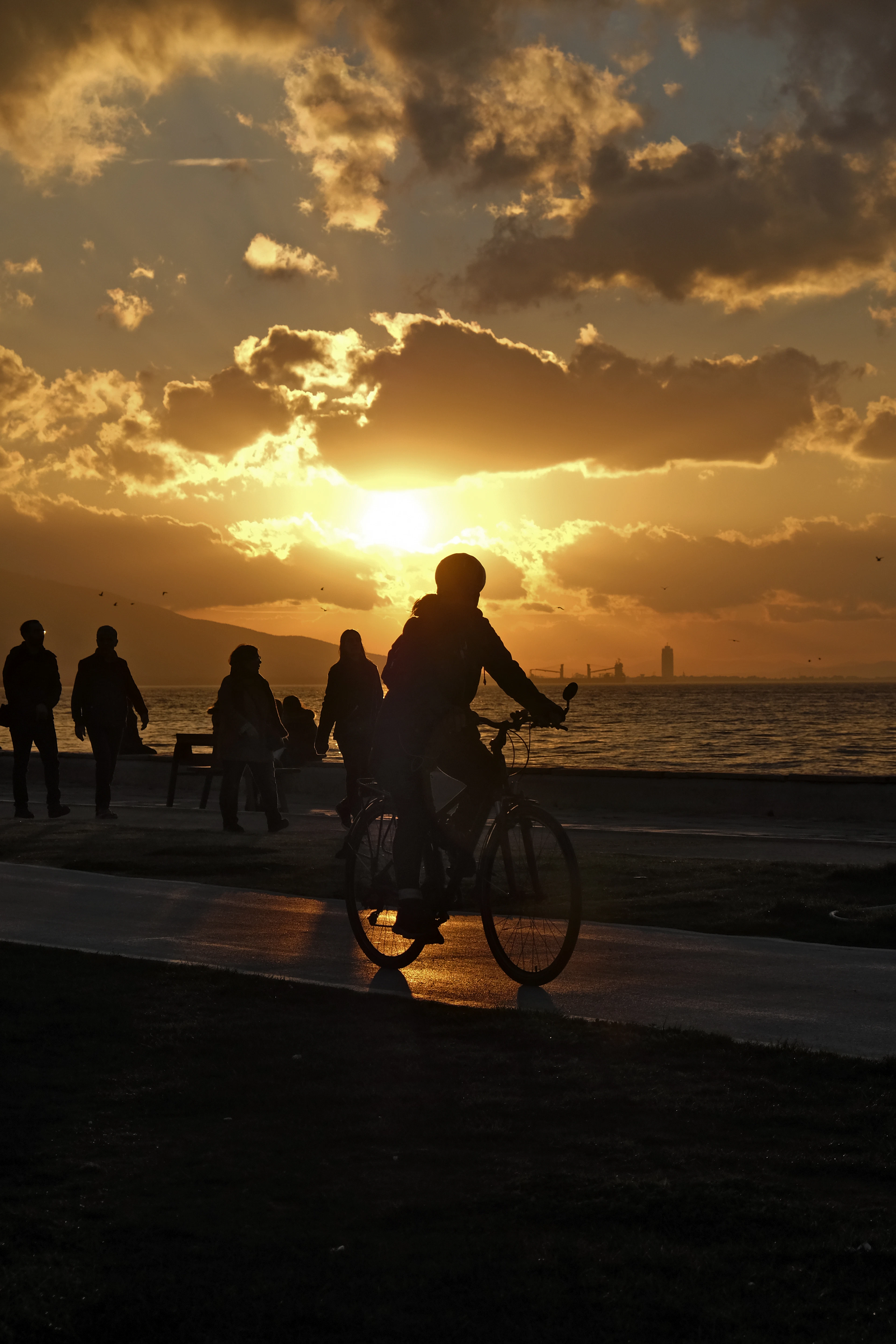Walkers and a cyclist in silhouette on Izmir's Kordon promenade as the sun drops behind the hills across the Gulf of Izmir, banked cloud burning orange above the water and the wet path catching the light
