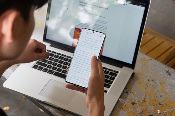 A person is sitting at a table using both a smartphone and a laptop. The phone displays a webpage with text, while the laptop screen also shows a similar webpage. Part of the table surface is visible, which appears to have a decorative pattern.