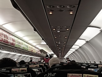 A view of the interior of an airplane cabin showing the overhead compartments, illuminated with soft lighting. Passengers are seated with a few people standing or moving in the aisle. Advertisements are visible on the overhead bins, and seat covers feature promotional text.