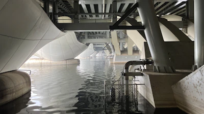 A view beneath a modern geometric bridge featuring large white and gray structural elements. The architecture exhibits curves and angles, creating a dynamic overpass above a body of still water. The reflections on the water emphasize the symmetry and complexity of the engineering design.