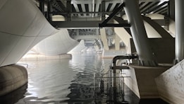 A view beneath a modern geometric bridge featuring large white and gray structural elements. The architecture exhibits curves and angles, creating a dynamic overpass above a body of still water. The reflections on the water emphasize the symmetry and complexity of the engineering design.