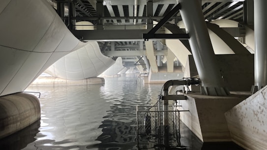 A view beneath a modern geometric bridge featuring large white and gray structural elements. The architecture exhibits curves and angles, creating a dynamic overpass above a body of still water. The reflections on the water emphasize the symmetry and complexity of the engineering design.