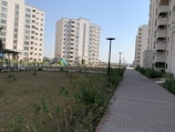 A residential complex with several mid-rise apartment buildings. The buildings are modern, featuring multiple balconies and large windows. A landscaped garden with a grassy area, plants, and trees is present beside a paved walkway. Streetlights line the walkway, and a colorful playground is visible in the background.