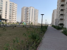 A residential complex with several mid-rise apartment buildings. The buildings are modern, featuring multiple balconies and large windows. A landscaped garden with a grassy area, plants, and trees is present beside a paved walkway. Streetlights line the walkway, and a colorful playground is visible in the background.