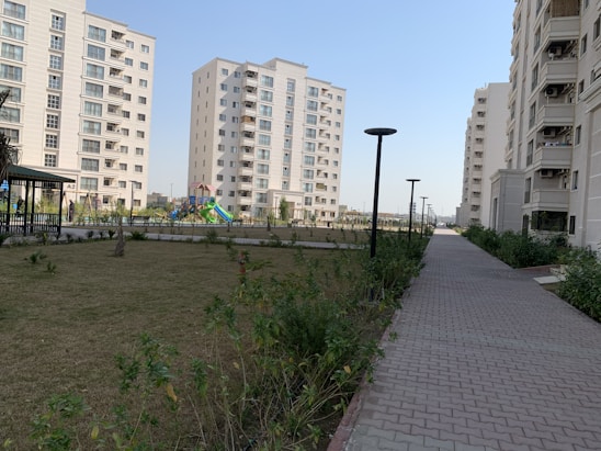 A residential complex with several mid-rise apartment buildings. The buildings are modern, featuring multiple balconies and large windows. A landscaped garden with a grassy area, plants, and trees is present beside a paved walkway. Streetlights line the walkway, and a colorful playground is visible in the background.