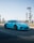 A vibrant blue sports car is driving on a road, surrounded by a landscape with sparse trees lining the roadside. The car features distinctive aerodynamic components and a sleek design, indicative of high performance.
