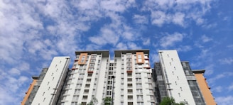 A high-rise residential building with modern architecture reaches towards a partly cloudy blue sky. The building features a combination of white and orange segments with numerous windows, and some parts have green foliage at the base.